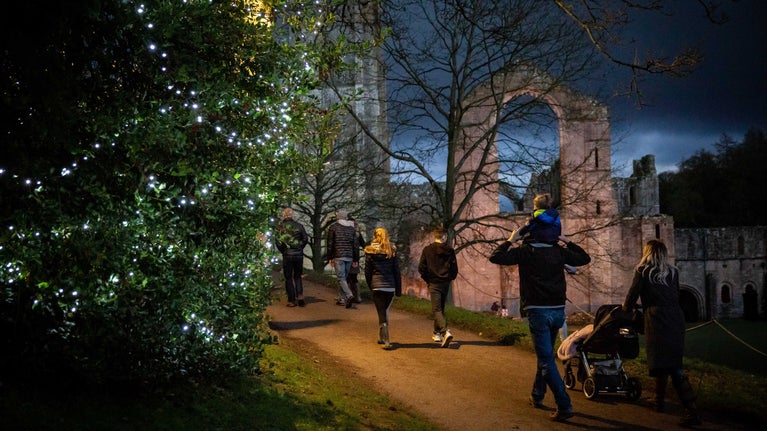 Visitors walking past the abbey as dusk starts to fall with twinkling lights filling the trees.